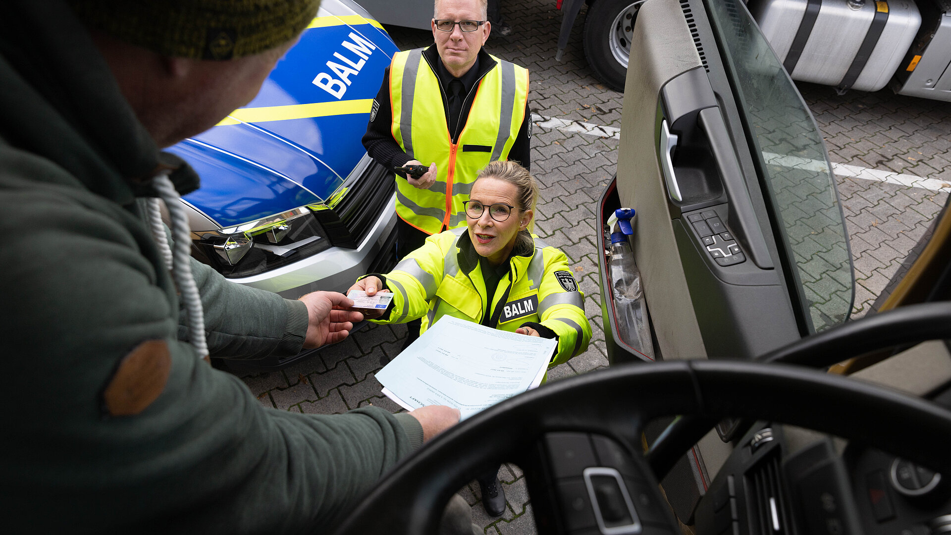 Ein Polizist übergibt einer Frau ein Dokument, während ein weiterer Polizist neben einem Fahrzeug steht