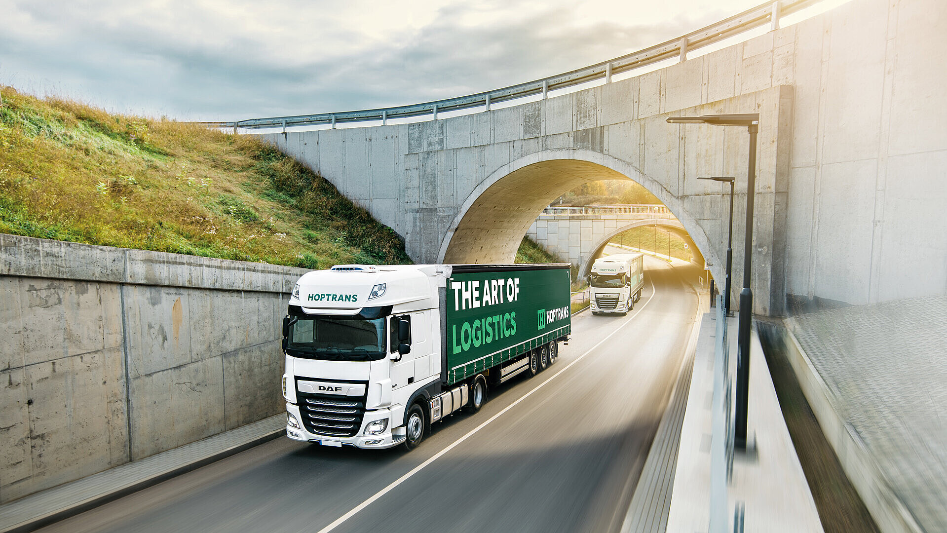 Ein weißer DAF-LKW fährt auf einer Straße unter einem Tunnel hindurch, während ein weiterer LKW im Hintergrund fährt